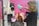 A hairdresser using a green hairdryer is drying the hair of a seated client in a salon with pink walls. Various salon tools and equipment are visible in the background.