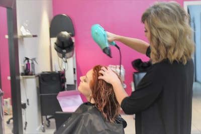 A hairdresser using a green hairdryer is drying the hair of a seated client in a salon with pink walls. Various salon tools and equipment are visible in the background.