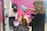 A hairdresser using a green hairdryer is drying the hair of a seated client in a salon with pink walls. Various salon tools and equipment are visible in the background.