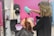 A hairdresser using a green hairdryer is drying the hair of a seated client in a salon with pink walls. Various salon tools and equipment are visible in the background.