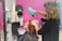 A hairdresser using a green hairdryer is drying the hair of a seated client in a salon with pink walls. Various salon tools and equipment are visible in the background.