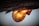 A close-up of a droplet of amber resin hanging from a tree branch. The resin is glowing with warm colors ranging from yellow to orange and red, reflecting light and creating a striking contrast against the darker background.