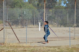 A child wearing a blue camouflage hoodie is preparing to throw a javelin on a fenced-off area with a rustic, aged look. The background includes sparse vegetation and a worn-out playing surface.
