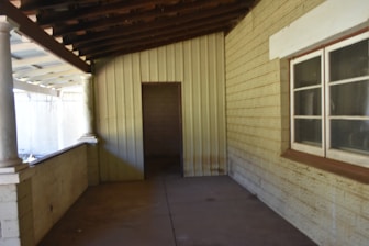 A covered porch area with a wooden ceiling and exposed beams. The walls are constructed with corrugated metal and textured blocks, featuring a window with multiple panes and a door leading to another space. Columns support the roof, and the floor appears to be concrete.