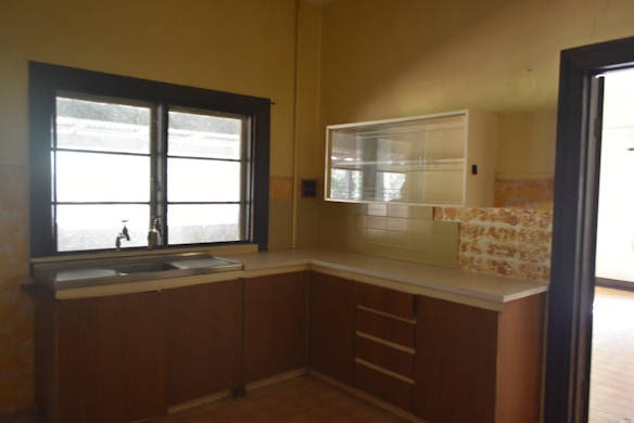 A kitchen with brown cabinets and a stainless steel sink under a large window. There is a glass-fronted cabinet above the counter, and the walls have a yellowish tone with some patterned tiles.
