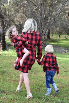 A woman with blonde hair is walking through a grassy area while holding a young child in her arms. Another child is holding the woman's hand. All three are wearing matching red and black plaid outfits. The background consists of trees and greenery, creating a natural and serene environment.