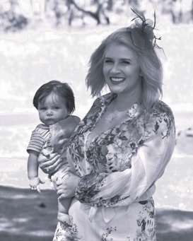 A woman smiling warmly, wearing a floral dress with long sleeves and a decorative fascinator in her hair, holding a baby dressed in overalls with a striped shirt. The background is blurred, and the scene appears to be outdoors with natural light.