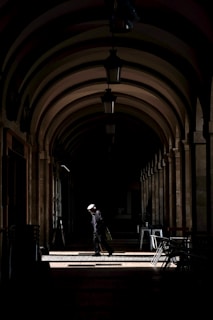 An elderly person wearing a hat and carrying a bag walks through a dimly lit, arched corridor. A single beam of sunlight highlights the individual, creating a strong contrast against the surrounding shadows. The arched ceiling and symmetrical columns add a sense of depth and architectural elegance.