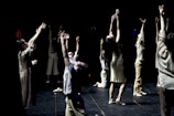 A group of dancers perform onstage with their arms raised towards the ceiling. They are dressed in casual, neutral-colored clothing, and the backdrop is dark, highlighting their movements. The lighting creates dramatic shadows, emphasizing the expressive nature of the dance.