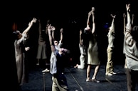 A group of dancers perform onstage with their arms raised towards the ceiling. They are dressed in casual, neutral-colored clothing, and the backdrop is dark, highlighting their movements. The lighting creates dramatic shadows, emphasizing the expressive nature of the dance.
