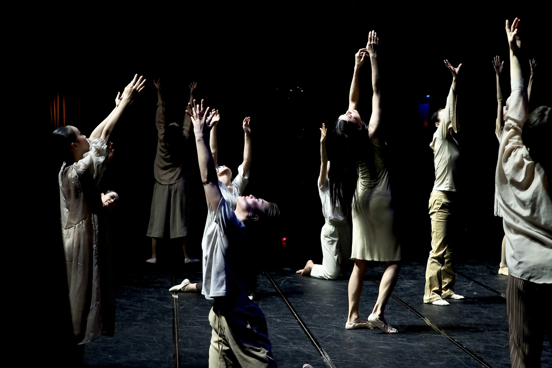 A group of dancers perform onstage with their arms raised towards the ceiling. They are dressed in casual, neutral-colored clothing, and the backdrop is dark, highlighting their movements. The lighting creates dramatic shadows, emphasizing the expressive nature of the dance.