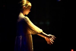 A young person stands in a dark space, illuminated by a soft, warm light, highlighting their features and casting delicate shadows. The individual appears to be deeply focused, with their hands slightly extended forward, creating an artistic and serene atmosphere.