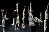 A group of individuals is engaged in a dance performance, each extending their arms upwards in a dramatic fashion. They are dressed in light-colored, flowing attire and are positioned on a dimly lit stage with a dark background that emphasizes their movements.