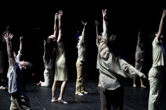 A group of individuals is engaged in a dance performance, each extending their arms upwards in a dramatic fashion. They are dressed in light-colored, flowing attire and are positioned on a dimly lit stage with a dark background that emphasizes their movements.