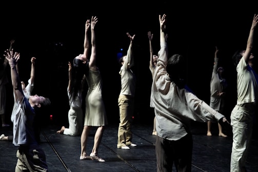 A group of individuals is engaged in a dance performance, each extending their arms upwards in a dramatic fashion. They are dressed in light-colored, flowing attire and are positioned on a dimly lit stage with a dark background that emphasizes their movements.