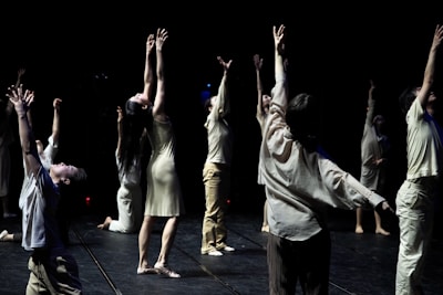 A group of individuals is engaged in a dance performance, each extending their arms upwards in a dramatic fashion. They are dressed in light-colored, flowing attire and are positioned on a dimly lit stage with a dark background that emphasizes their movements.