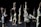 A group of individuals is engaged in a dance performance, each extending their arms upwards in a dramatic fashion. They are dressed in light-colored, flowing attire and are positioned on a dimly lit stage with a dark background that emphasizes their movements.