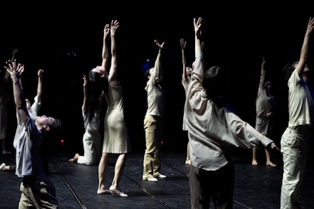 A group of individuals is engaged in a dance performance, each extending their arms upwards in a dramatic fashion. They are dressed in light-colored, flowing attire and are positioned on a dimly lit stage with a dark background that emphasizes their movements.