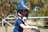 A young child wearing a large, black and white motocross helmet is sitting on a bicycle or small dirt bike. The child is dressed in a blue t-shirt and is positioned in front of a metal gate, with a rural background that includes trees and a grassy area.