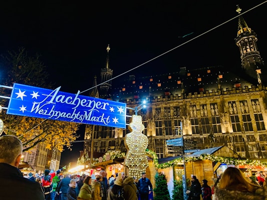 A vibrant Christmas market bustling with people under a dark night sky. The image features a large historic building illuminated by warm lights. A blue sign with white stars reads 'Aachener Weihnachtsmarkt.' Stalls decorated with festive lights and garlands line the market area. A mix of bare trees and those with twinkling lights add to the festive atmosphere.