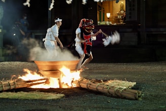 A traditional Japanese ceremony is taking place outdoors at night. Two participants, one wearing a red dragon mask and the other in white attire, are performing a ritual around a fire pit. The scene is illuminated by the flames, creating a dramatic and mystical atmosphere. The background shows part of a building, possibly a shrine, with soft ambient lighting.