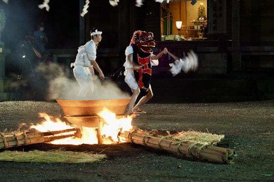 A traditional Japanese ceremony is taking place outdoors at night. Two participants, one wearing a red dragon mask and the other in white attire, are performing a ritual around a fire pit. The scene is illuminated by the flames, creating a dramatic and mystical atmosphere. The background shows part of a building, possibly a shrine, with soft ambient lighting.