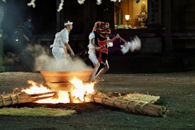 A traditional Japanese ceremony is taking place outdoors at night. Two participants, one wearing a red dragon mask and the other in white attire, are performing a ritual around a fire pit. The scene is illuminated by the flames, creating a dramatic and mystical atmosphere. The background shows part of a building, possibly a shrine, with soft ambient lighting.