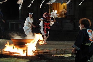 A traditional Japanese festival scene with participants dressed in ceremonial attire. One person wears a red and black costume, likely representing a mythical creature, and holds ritual objects. Another person in white ceremonial clothing accompanies them. A fire burns in a large metal container, and there are onlookers in traditional dress nearby. The backdrop presents a shrine-like structure with traditional decorations and lighting, enhancing the cultural ambiance.