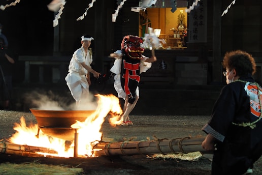 A traditional Japanese festival scene with participants dressed in ceremonial attire. One person wears a red and black costume, likely representing a mythical creature, and holds ritual objects. Another person in white ceremonial clothing accompanies them. A fire burns in a large metal container, and there are onlookers in traditional dress nearby. The backdrop presents a shrine-like structure with traditional decorations and lighting, enhancing the cultural ambiance.