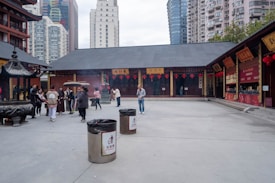 A courtyard with traditional architecture and red lanterns hanging from the buildings. Several people are standing in small groups, and there are large incense burners in the foreground. High-rise buildings are visible in the background, blending modern and traditional elements.