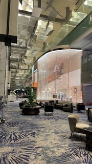 A modern indoor space featuring sleek, metallic architectural elements with a patterned wall incasing an upper-level area. The setting is adorned with contemporary furniture, including cushioned chairs, sofas, and coffee tables arranged on a patterned blue carpet. A large plant provides a touch of greenery amid the metallic and glass structures, and the ceiling has an intricate, geometric design with sections suspended above.