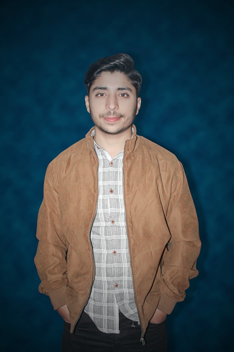 A young man stands against a deep blue background, wearing a tan jacket over a plaid shirt. His hands are in his jacket pockets, and he looks directly at the camera with a neutral expression. His dark hair is styled neatly.