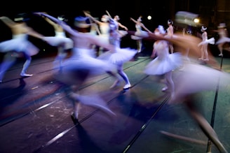 Ballerinas dressed in white tutus are gracefully performing on stage, with blurred motion capturing the dynamism of their dance. The stage is dimly lit with spotlights adding a dramatic effect, and their synchronized movement creates a sense of fluidity and elegance.