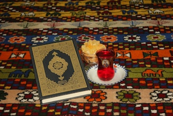 A richly patterned carpet serves as the backdrop for a religious book with intricate golden designs on the cover. A glass of red liquid sits on a decorative saucer, accompanied by a bowl of light-colored snacks, possibly candied or dried fruit. The carpet features vibrant colors and motifs, including animals and flowers.