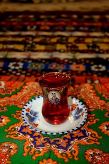 A glass of red tea is placed on a decorative white saucer with blue floral patterns. The tablecloth beneath features elaborate patterns with vibrant orange, green, blue, and red colors, adding an intricate and exotic feel.
