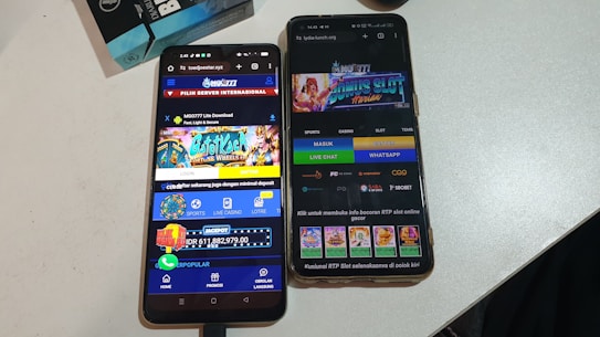 Two smartphones are displayed on a white surface, both showing gambling websites with colorful graphics, text, and various options for logging in and accessing games. The screens are bright and filled with promotional content related to casino and slot games.