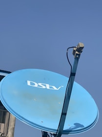 A blue satellite dish with the DSTV logo mounted on a pole. The dish is facing upwards against a clear blue sky, with a cable attached to the arm supporting the dish.
