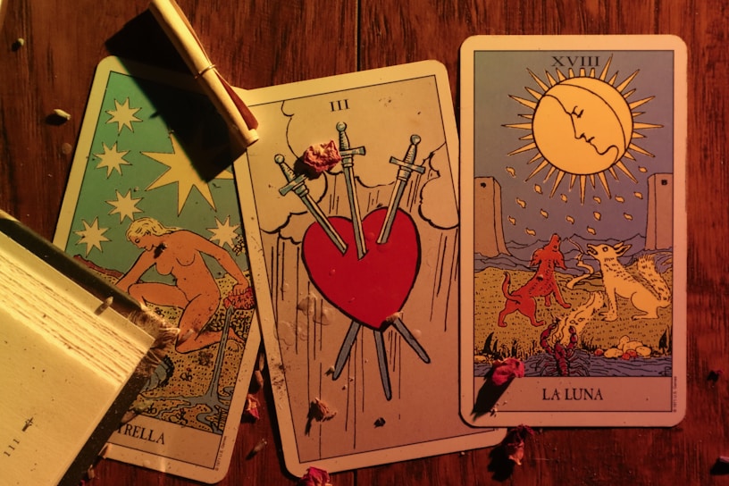 Three tarot cards are laid out on a wooden surface. The left card features a nude figure pouring water, surrounded by stars, and is labeled 'LA STELLA.' The center card depicts a heart pierced by three swords, symbolizing heartbreak or sorrow, and is labeled with the Roman numeral III. The right card shows a landscape with a large sun or moon, two animals, and two towers in the background. It is labeled 'LA LUNA' with the Roman numeral XVIII. A partially burned scroll is placed on top of the leftmost card, and there are scattered dried flowers on the table.