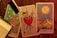 Three tarot cards are laid out on a wooden surface. The left card features a nude figure pouring water, surrounded by stars, and is labeled 'LA STELLA.' The center card depicts a heart pierced by three swords, symbolizing heartbreak or sorrow, and is labeled with the Roman numeral III. The right card shows a landscape with a large sun or moon, two animals, and two towers in the background. It is labeled 'LA LUNA' with the Roman numeral XVIII. A partially burned scroll is placed on top of the leftmost card, and there are scattered dried flowers on the table.