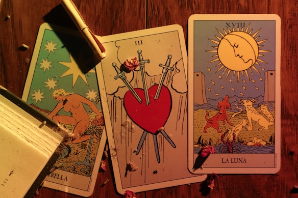 Three tarot cards are laid out on a wooden surface. The left card features a nude figure pouring water, surrounded by stars, and is labeled 'LA STELLA.' The center card depicts a heart pierced by three swords, symbolizing heartbreak or sorrow, and is labeled with the Roman numeral III. The right card shows a landscape with a large sun or moon, two animals, and two towers in the background. It is labeled 'LA LUNA' with the Roman numeral XVIII. A partially burned scroll is placed on top of the leftmost card, and there are scattered dried flowers on the table.