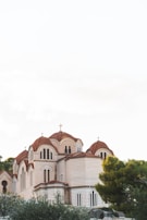 A traditional Byzantine-style church with multiple small domes and crosses on top, featuring intricate brickwork and arched windows. The structure is surrounded by lush greenery and set against a clear sky.