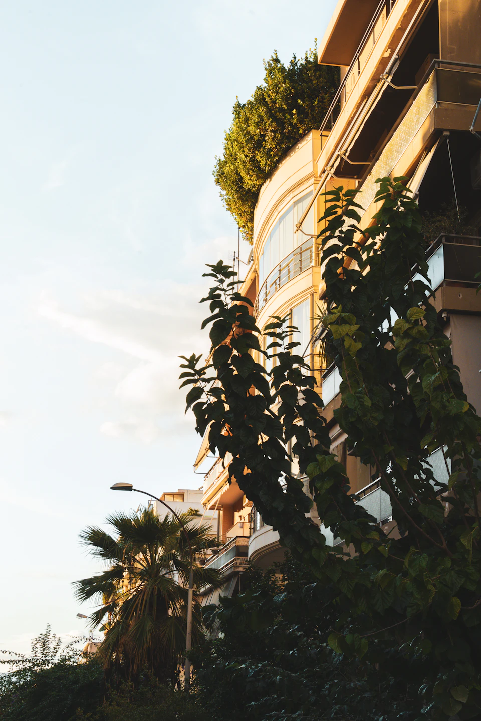 The image depicts a tall, narrow building with a curved facade and balconies on all sides. The building is situated amidst lush greenery, including palm trees that add to its tropical ambiance. A street lamp stands nearby, providing illumination for the area. The perspective of the photo is taken from below, looking up at the building, which enhances its imposing presence against the backdrop of a clear blue sky with fluffy white clouds.

The image also contains some text, but it is not legible and does not provide any additional information about the scene or the building's history.