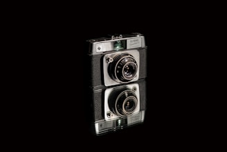 A vintage camera with a black and silver body, featuring intricate lens details, stands prominently against a dark background. The camera is artistically reflected on a shiny surface beneath it.