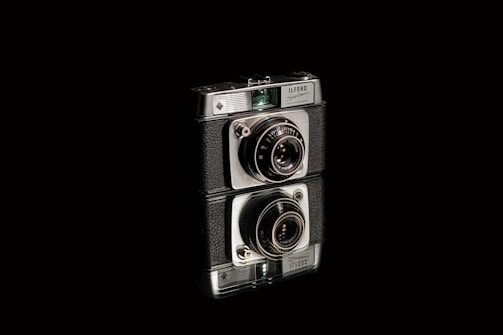 A vintage camera with a black and silver body, featuring intricate lens details, stands prominently against a dark background. The camera is artistically reflected on a shiny surface beneath it.