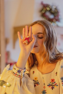 A person wearing a cream-colored blouse with colorful floral embroidery holds a small angel ornament with a red bow in front of their face. The background is softly blurred, with a wreath visible, creating a festive atmosphere.