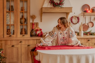A person in a floral embroidered blouse appears to be preparing a round dining table with a red tablecloth, set in a cozy kitchen featuring wooden cabinets and rustic kitchenware. Holiday decorations such as wreaths and lit candles add a festive touch.