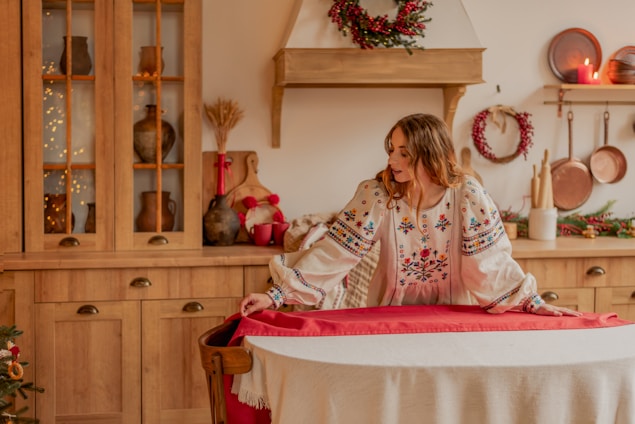 A person in a floral embroidered blouse appears to be preparing a round dining table with a red tablecloth, set in a cozy kitchen featuring wooden cabinets and rustic kitchenware. Holiday decorations such as wreaths and lit candles add a festive touch.