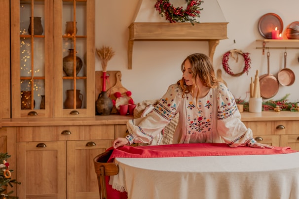 A person in a floral embroidered blouse appears to be preparing a round dining table with a red tablecloth, set in a cozy kitchen featuring wooden cabinets and rustic kitchenware. Holiday decorations such as wreaths and lit candles add a festive touch.
