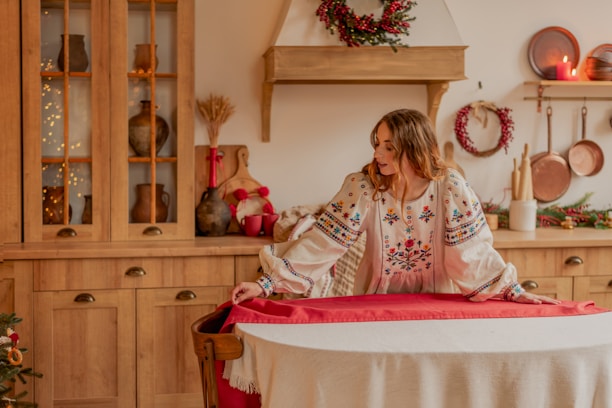 A person in a floral embroidered blouse appears to be preparing a round dining table with a red tablecloth, set in a cozy kitchen featuring wooden cabinets and rustic kitchenware. Holiday decorations such as wreaths and lit candles add a festive touch.