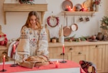 A woman wearing a traditional embroidered dress is placing a freshly baked loaf of bread on a table covered with a red cloth. The room is decorated for the holiday season with evergreen arrangements and red candles. Copper pans and other utensils hang on the wall, adding a rustic charm.